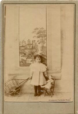 España, retrato de una niña con una muñeca en su sillita y un carrito de juguete, situada delante de una pintura. Fotógrafo desconocido.