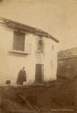 España, vivienda rural en un lugar desconocido con una capillita sobre la puerta principal y un hombre con una capa. Fotógrafo desconocido.